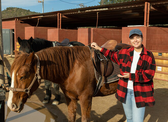 Portrait of smiling young European woman adjusting saddle of horse before horseback ride in country...