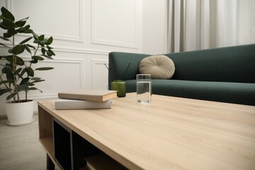 Books, glass of water and reed diffuser on wooden coffee table near sofa in living room, closeup