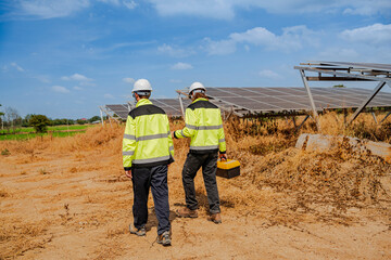 Fototapeta premium Two technicians in high-visibility jackets and helmets walk through a solar farm, carrying tools and a laptop, inspecting photovoltaic panels surrounded by dry grass under a clear sky.