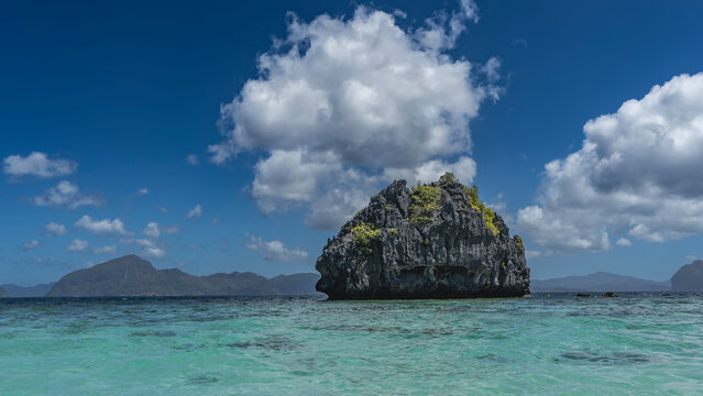A rocky island in a turquoise ocean. There is green vegetation on the steep furrowed slopes. Mountains in the distance. Blue sky, clouds. Philippines. Palawan. Bacuit Bay.