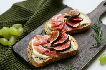 Wooden board of delicious fig bruschetta with prosciutto and ricotta cheese on white background,...