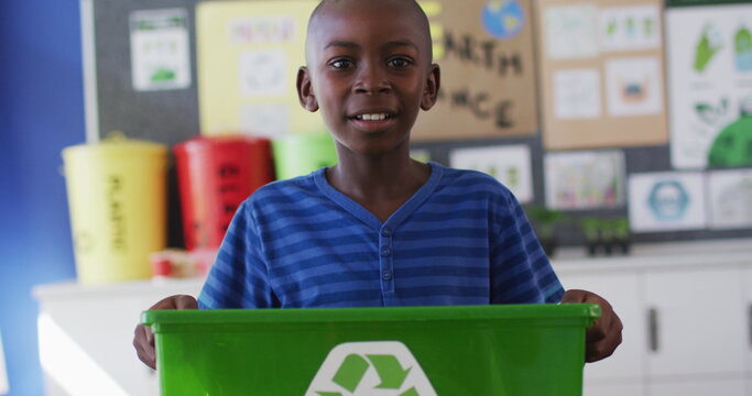 African american schoolboy smiling, holding recycling bin, standing in classroom