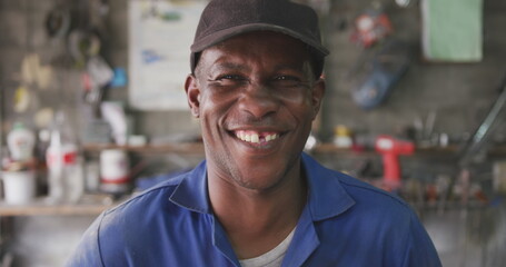 Fototapeta premium Portrait of a happy African male panel beater in a township workshop, wearing a cap, looking at came