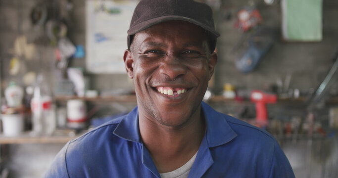 Portrait of a happy African male panel beater in a township workshop, wearing a cap, looking at came