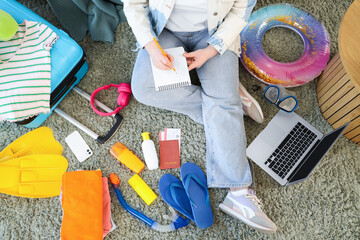 Young woman making list with travel accessories on carpet at home, top view