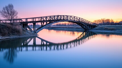 Obraz premium A serene wooden bridge arches over calm waters at sunset, reflecting vibrant colors in the sky, creating a tranquil landscape.
