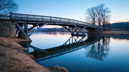 A tranquil wooden bridge spans a calm river, reflecting the twilight sky and surrounded by bare trees.