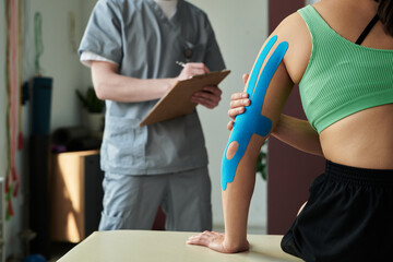 Woman receiving treatment from physiotherapist in medical clinic. Therapist observing and taking notes while patient with kinesiology tape on arm sits on examination table