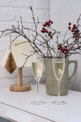 Glasses of champagne with Christmas dry tree branches with blurred lights and red berries, christmas tree on table in living room.