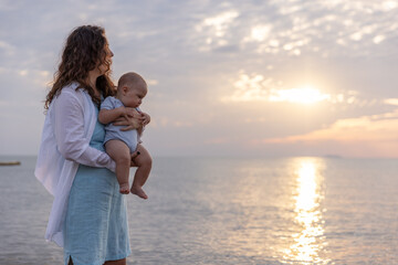 Mother Holding Baby at Sunset by the Sea, Watching the Reflection of Sunlight on the Water, Creating a Peaceful and Emotional Moment Between Mother and Child at the Beach