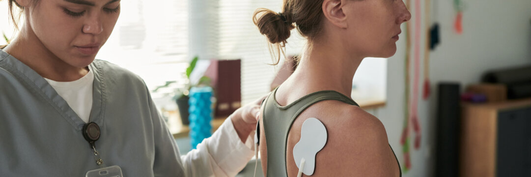 Medical professional placing electrode pad on patient's upper back in clinic environment. Patient with shoulder exposed, receiving treatment from healthcare worker