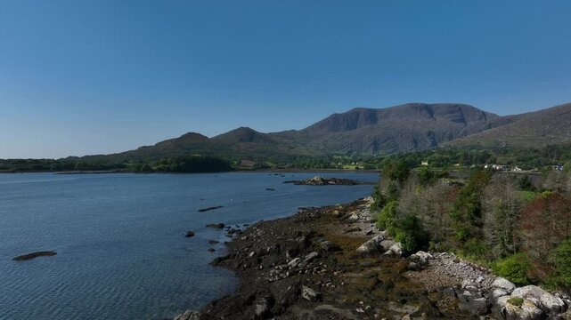 Adrigole Harbour, Beara Peninsula, County Cork, Ireland, September 2024. Drone pushes forwards along the craggy rocky coastline with Coomgira and Caha Mountains in the distance under a clear blue sky.