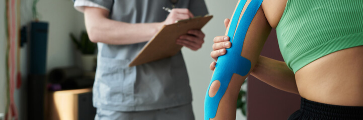 Therapist reviewing athletic tape application on patient during therapy session for muscle pain relief. Therapist and patient engaged in detailed examination of tape placement