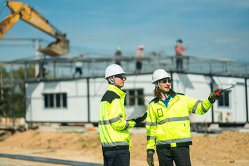 Two engineers wearing high visibility jackets and helmets discussing construction progress at a site, one pointing towards workers and machinery, emphasizing teamwork, safety, and planning in