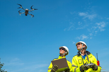 Two engineers in high visibility jackets and helmets observing a drone flying over a construction...