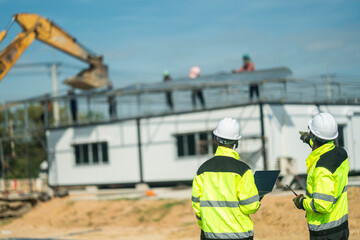 Two engineers wearing high visibility jackets and helmets discussing construction progress at a...