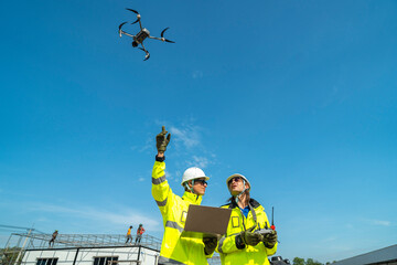Two engineers in high visibility jackets and helmets observing a drone flying over a construction...
