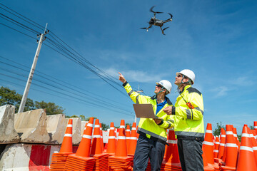 Two engineers in high visibility jackets and helmets operate a drone using a laptop amidst a stack...