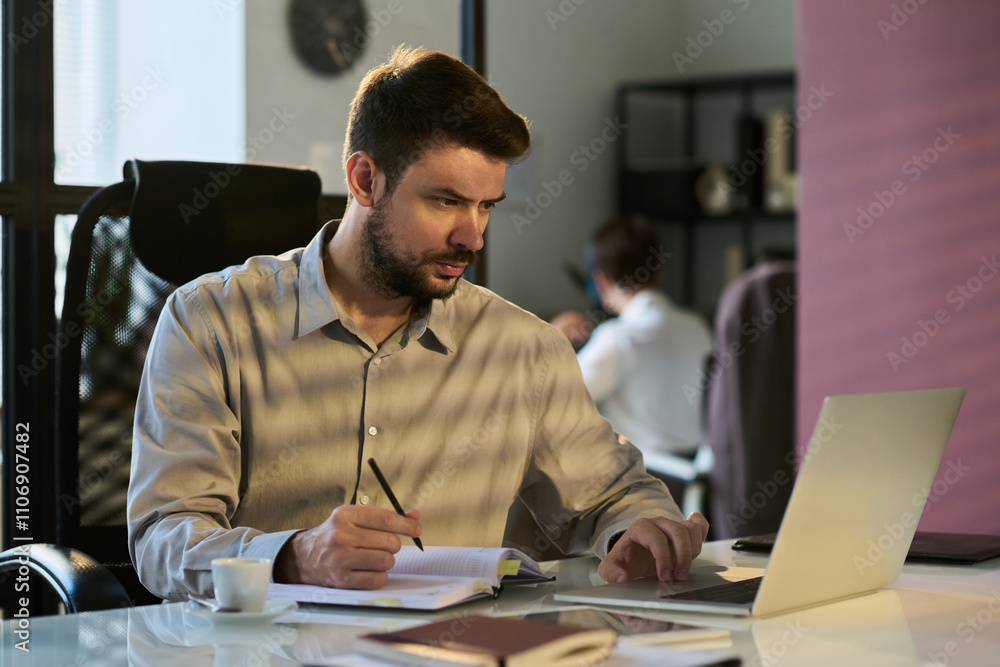 Poster Young serious male white collar worker looking at laptop screen while analyzing data and taking notes in notepad