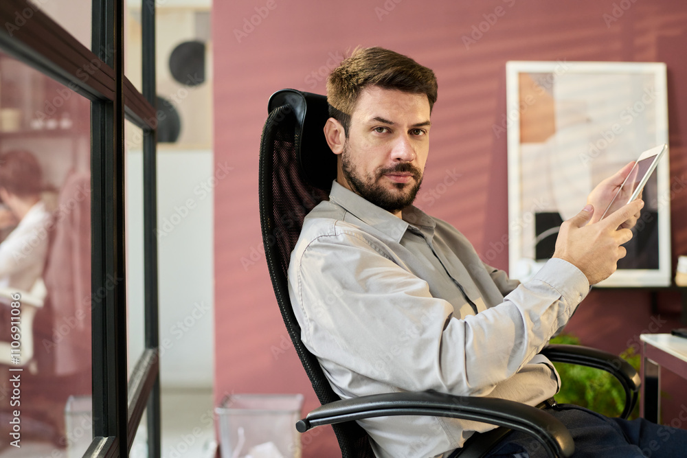 Poster young confident businessman with mobile gadget looking at camera while sitting in armchair by his wo
