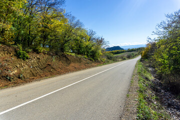 An asphalt road with a white dividing line passes through trees and bushes with autumn leaves. Hills and clouds are in the background