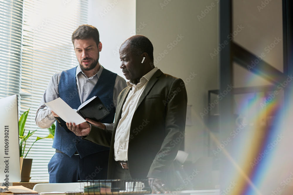 Poster young male economist with open notepad explaining data on paper document held by mature african amer