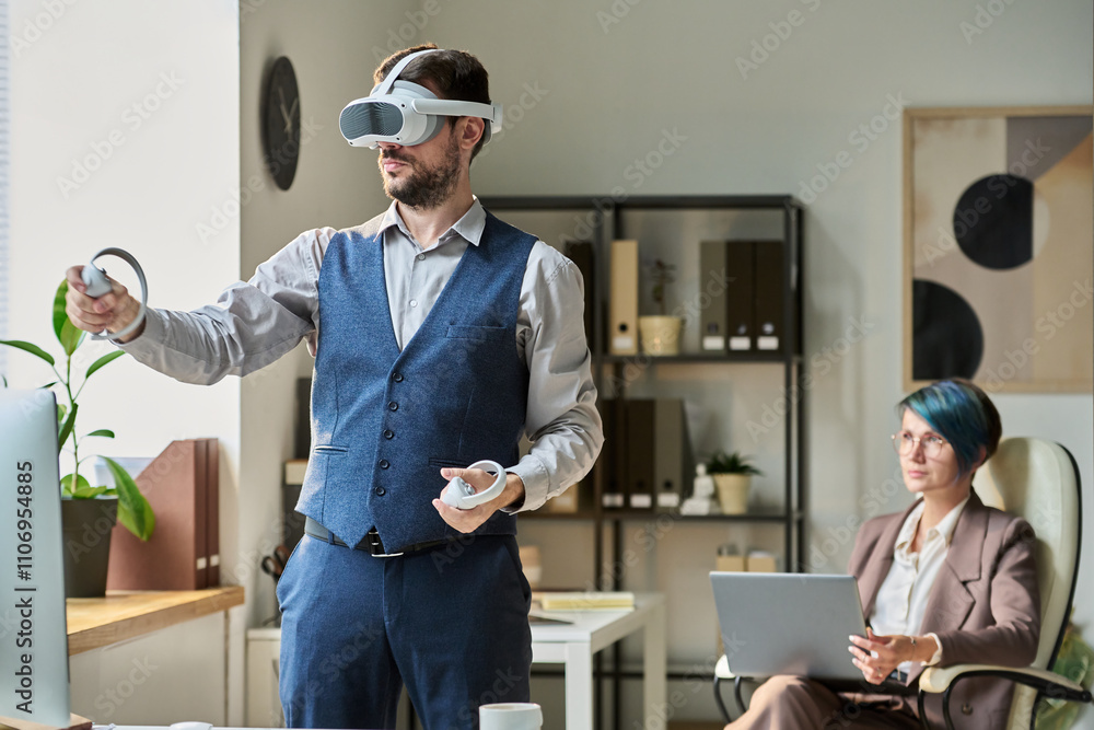 Poster Young modern businessman with VR headset using controllers while making presentation of new project against colleague