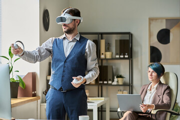 Young modern businessman with VR headset using controllers while making presentation of new project against colleague