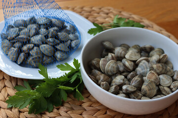 Many littleneck fresh clams in a white bowl with parsley leaves on wooden table