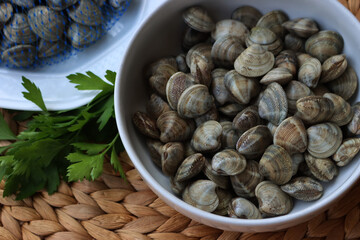 Many littleneck fresh clams in a white bowl with parsley leaves on wooden table