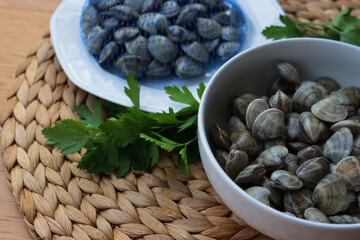 Many littleneck fresh clams in a white bowl with parsley leaves on wooden table