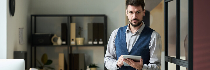 Header of young serious businessman using tablet and looking at camera while standing in coworking space with desks and shelves