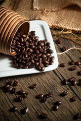 A rustic wooden cup spills its contents of dark roasted coffee beans onto a pristine white plate. The beans are scattered haphazardly across a weathered wooden surface.