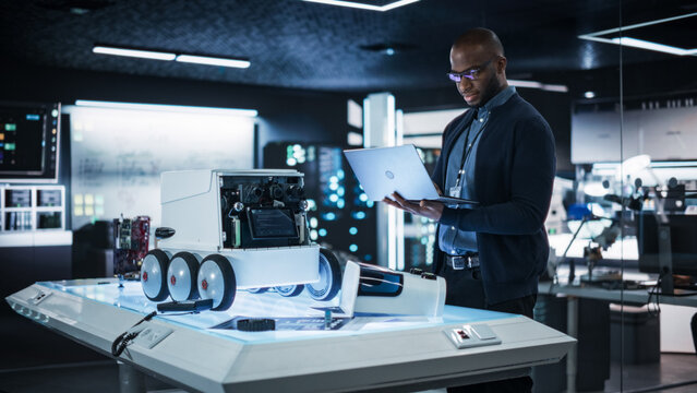 Portrait of an African Engineer Using a Laptop Computer to Write a Report on Delivery Robot Prototype in a Research Laboratory. Young Black Quality Assurance Specialist Reporting Bugs in Code