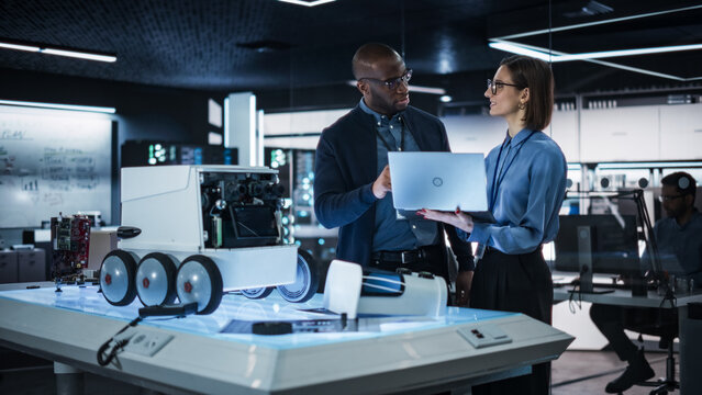 Colleagues Analysing Data Using Laptop Computer and Brainstorming for a Project: Portrait of a Team of Two Engineers Collaborating on Developing a Robot for Deliveries in a Manufacturing Facility
