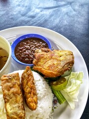 Nasi Ayam Penyet with fried chicken, fried tofu, tempe, vegetables, lime, soup, and sambal belacan on dark background. Famous traditional Indonesian food.