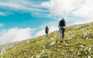 Two hikers walking uphill on a rocky, grassy terrain under a bright blue sky with scattered clouds....