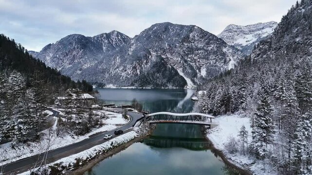 Drone view of Plansee lake in Austria, featuring a snowy bridge, mountains, and a winding road in winter