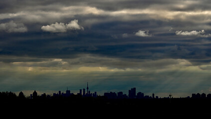 A sky covered with dark clouds but with a hint of sunshine, paired with the silhouette of the city of Toronto and the CN Tower