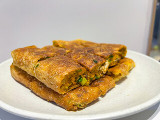A platter of golden-brown martabak telur, a traditional Indonesian street food, is presented on a white plate. Each slice reveals a savory filling of egg, meat, chives, and other ingredients