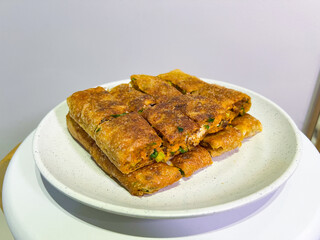 A platter of golden-brown martabak telur, a traditional Indonesian street food, is presented on a white plate. Each slice reveals a savory filling of egg, meat, chives, and other ingredients