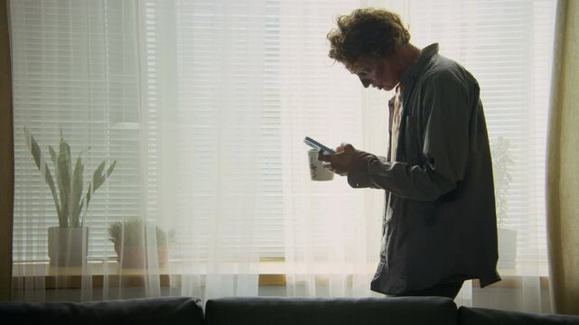 Medium lockdown shot of lethargic undead man with decomposing flesh, in bloody clothing stumbling by window at home, reading messages or social media on smartphone, drinking coffee, walking off