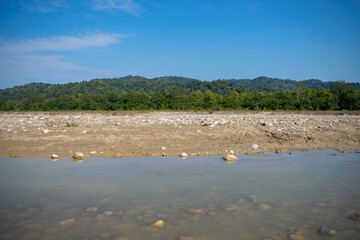 A tranquil riverbank with clear water, scattered rocks, and a sandy shore set against a backdrop of dense green forest and blue skies. The serene landscape is perfect for nature and outdoor themes.
