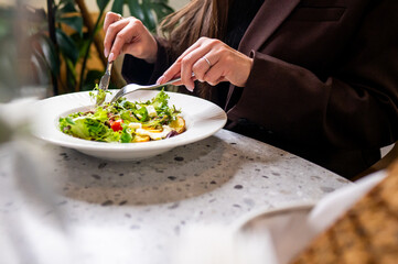 A close-up of a person enjoying a healthy meal. Hands with utensils are seen serving a colorful salad on a stylish plate, surrounded by an elegant dining setting. Fresh ingredients are highlighted.