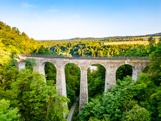 Fototapeta premium The Zampach stone railway bridge spans lush trees, showcasing impressive stone arches against a clear sky, set in the scenic Czech countryside.