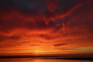 Obraz premium Dramatic sunset over calm lake with fiery red and orange clouds illuminating the sky.