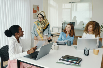 Fototapeta premium Diverse group of women discussing work while seated around table in modern office. Collaboration and teamwork are evident. Various devices and documents are spread across the table