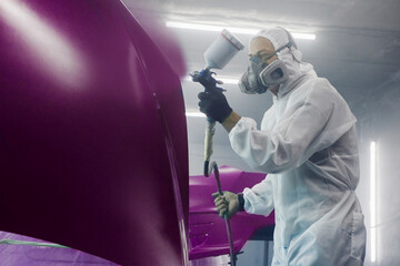 Professional spray painter in protective mask and clothes spraying car hood of vehicle part to...