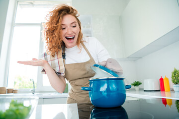 Charming redhead woman joyfully cooking a dish in cozy kitchen under natural daylight