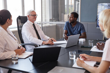 Experienced senior male clinician discussing anamnesis of patients disease with diverse group of colleagues sitting at meeting table while planning treatment at medical office